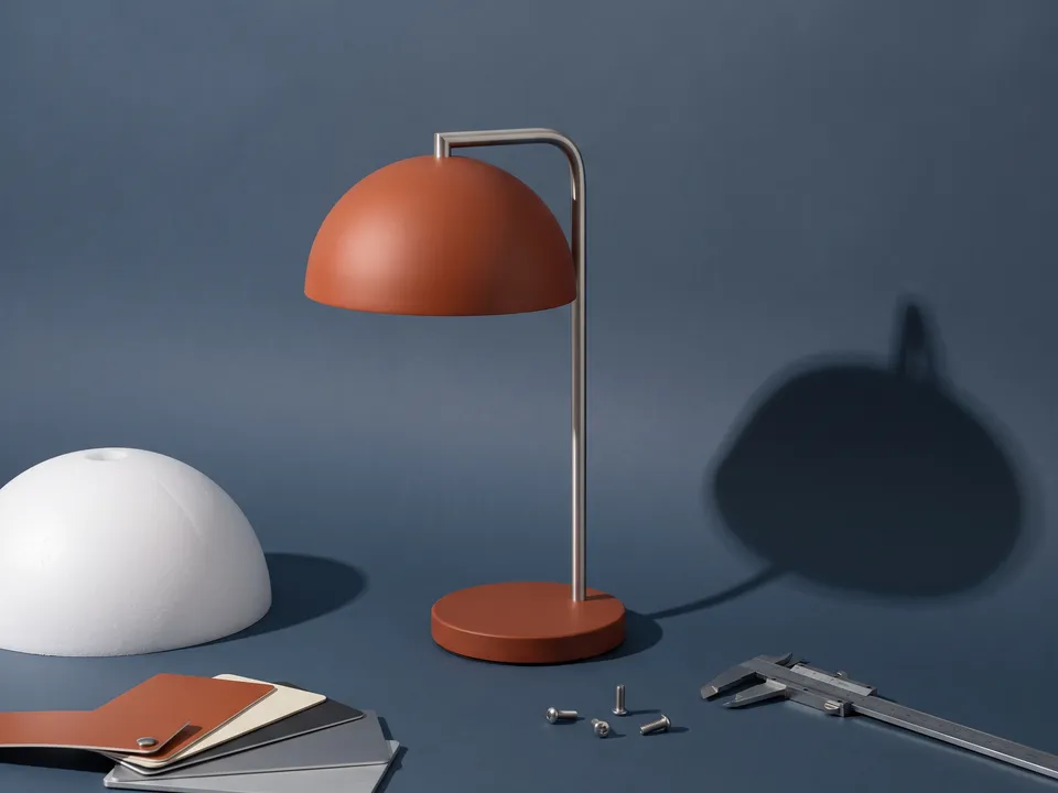 A desk lamp with a burnt-orange dome shade on slate blue, beside a white foam prototype of the shade, material chips and a caliper