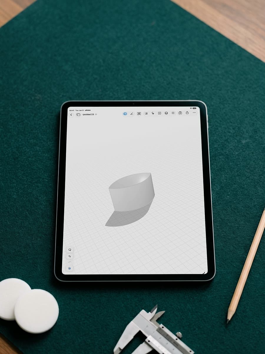 An iPad on a green felt mat showing the curved 3D solid left where two shapes overlap, beside white discs, calipers and a pencil